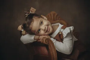Bambina appoggiata su una poltrona d'epoca durante un servizio fotografico a Roma, sorriso spontaneo, sfondo caldo dal tocco pittorico